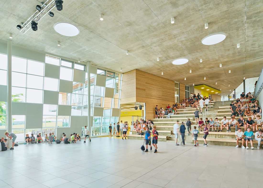 <p><span>The assembly hall is the centrepiece of the school. Its expansive, open steps and stepped seating offer plenty of options for sitting and playing. Slim, round Modul R Project luminaires from Nimbus create a fascinating contrast to the glass facade, which is lent a playful vibrancy by alternating rectangular window surfaces and white acoustic panels. Photo: David Matthiessen</span><span></span></p>